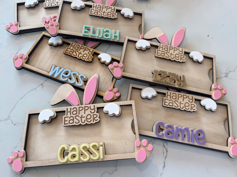 May include: A set of six wooden Easter bunny name tags with pink bunny ears, white clouds, and pink paw prints. Each tag has a different name and the text "Happy Easter".