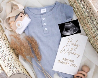 May include: A white card with the text "Baby Williams Arriving January 2022" in gold lettering. The card is surrounded by a blue baby onesie, a wooden camera, a wooden rattle, and dried grass.