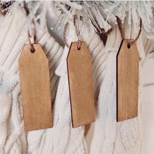 May include: Three blank wooden tags with a natural wood grain hang from twine strings. The tags are rectangular with a slightly tapered shape and a hole at the top. They are suspended against a backdrop of white knitted fabric and frosted greenery.