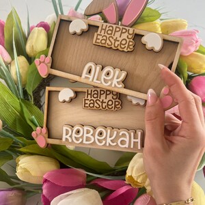 May include: Two wooden Easter bunny signs with pink ears and white paw prints. The top sign says "Happy Easter" and "Alek". The bottom sign says "Happy Easter" and "Rebekah".