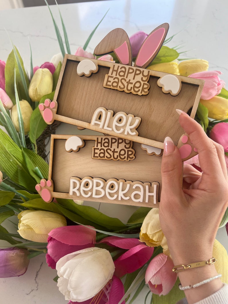 May include: Two wooden Easter bunny signs with pink ears and white paw prints. The top sign says "Happy Easter" and "Alek". The bottom sign says "Happy Easter" and "Rebekah".