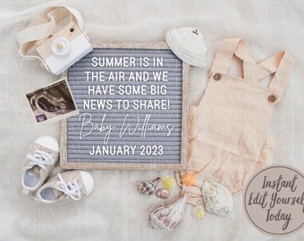 Op de afbeelding: Een grijs letterbord met de tekst "SUMMER IS IN THE AIR AND WE HAVE SOME BIG NEWS TO SHARE! Baby Williams JANUARY 2023". Er zijn schelpen, een paar babyschoentjes, een baby-romper en een speelgoedcamera op een wit oppervlak.