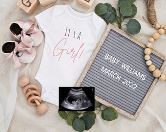 Op de afbeelding: Een wit rompertje met de tekst "It's a Girl!" in roze schrift. Een letterbord met de tekst "BABY WILLIAMS MARCH 2022". Een houten rammelaar, een paar roze babyschoentjes en een houten blok dat "GIRL" spelt. Een zwart-wit echografiebeeld.