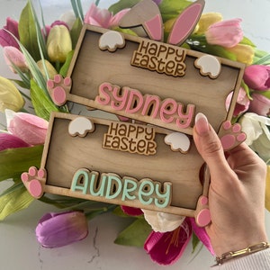 May include: Two wooden Easter bunny signs with pink bunny ears and paw prints. The top sign says "Happy Easter" and "Sydney". The bottom sign says "Happy Easter" and "Audrey".