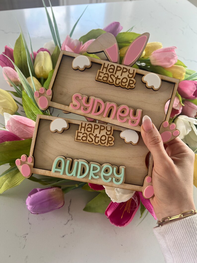 May include: Two wooden Easter bunny signs with pink bunny ears and paw prints. The top sign says "Happy Easter" and "Sydney". The bottom sign says "Happy Easter" and "Audrey".