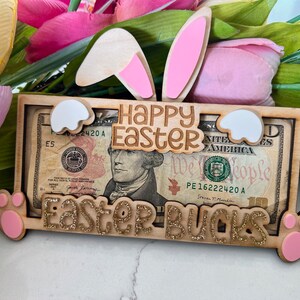 May include: A wooden Easter money holder with a bunny ear design. The holder has a slot for a dollar bill and says "Happy Easter" and "Easter Bucks".