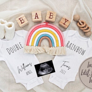 Puede incluir: Dos bodies blancos con el texto "Double Rainbow" y "Twins 2022" en ellos. Los bodies est&aacute;n colocados sobre un fondo de tela blanca con una decoraci&oacute;n de pared de arco&iacute;ris de hilo, bloques de madera que deletrean "BABY" y un sonajero de madera.