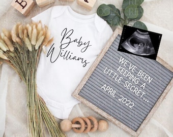 May include: A white onesie with the text "Baby Williams" in black script. A letter board with the text "We've been keeping a little secret... April 2022" is next to the onesie. There are wooden blocks spelling "BABY" and a wooden rattle.