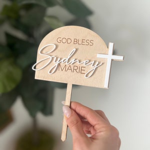 May include: A wooden cake topper with the words "GOD BLESS Sydney MARIE" in white script. A white cross is on the right side. The topper is held in front of a blurred green plant.