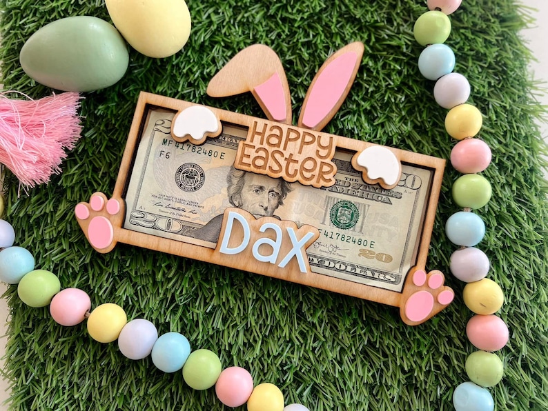 May include: A wooden bunny-shaped money holder with pink ears and a white paw print. The holder has a slot for a dollar bill and says "Happy Easter Dax" on it.