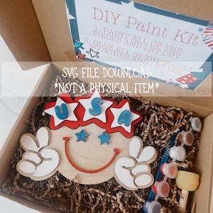 Puede incluir: Un kit de pintura DIY en una caja de cartón, con un diseño de madera con temática de EE. UU. en colores rojo, blanco y azul. El kit incluye botes de pintura, pinceles y una esponja. El texto en la caja dice "DIY Paint Kit" y "SVG FILE DOWNLOAD *NOT A PHYSICAL ITEM*".