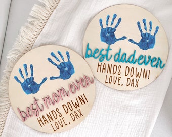 Könnte beinhalten: Zwei runde Holzschilder mit blauen Handabdrücken und dem Text "best dad ever HANDS DOWN! LOVE, DAX" und "best mom ever HANDS DOWN! LOVE, DAX" in rosa und blauen Buchstaben.