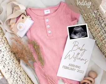 May include: A pink baby onesie with buttons, a wooden toy camera, a white card with the text "Baby Williams Arriving February 2022", a black and white ultrasound image, and a wooden rattle in a wicker basket.