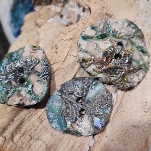 Peut inclure: Trois boutons en céramique faits à la main avec des textures et des couleurs uniques. Les boutons présentent un mélange de glaçures métalliques vertes, argentées et dorées sur une base beige. Chaque bouton a deux trous pour coudre.