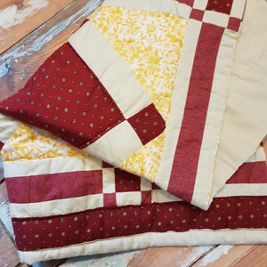 Op de afbeelding: Een patchworkdeken met een crèmekleurige achtergrond en rode en gele vierkanten. De deken is gemaakt van verschillende stoffen met verschillende patronen, waaronder bloemen- en geometrische patronen.