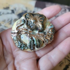 May include: A handcrafted, round ceramic button with a textured surface. The button features a raised, organic design with bronze and teal accents over a cream-colored base. The button is held in a hand.