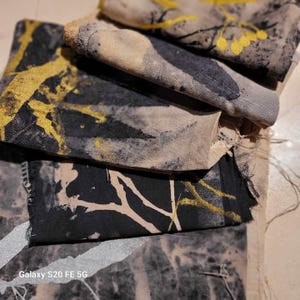 Könnte beinhalten: Ein Stapel aus vier Stoffquadraten mit einem abstrakten Design in Schwarz und Gold. Der Stoff ist hellbeige mit schwarzen und goldenen Farbstichen.