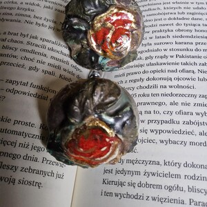 May include: Two handcrafted, round ornaments with a floral design. The ornaments feature a dark, textured exterior with a vibrant red and gold rose in the center. They are resting on the open pages of a book.
