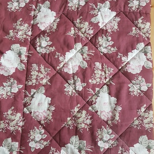 Puede incluir: Una manta acolchada de color burdeos con un patrón floral con rosas blancas y rosas. La manta tiene un patrón de acolchado en forma de diamante.