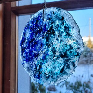 Könnte beinhalten: Ein runder, durchscheinender Glasschmuck mit strukturierter Oberfläche. Das Ornament zeigt eine Mischung aus Blau- und Türkistönen mit dunkleren blauen Akzenten. Es wird von einem dünnen Faden gehalten und hängt vor einem Fenster.