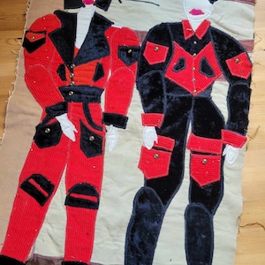 以下が含まれることがあります： 赤と黒の衣装を着た2人の様式化された人物を描いたテキスタイルアート。左側の人物は黒のアクセントが付いた赤いコーデュロイのスーツを着ており、右側の人物は赤いディテールが付いた黒のベルベットのスーツを着ています。「KLASA IIIA」の文字が見えます。