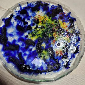Könnte beinhalten: Ein runder, klarer Glasuntersetzer mit einem farbenfrohen, abstrakten Design. Das Design zeigt eine Mischung aus blauen, schwarzen, gelben, grünen und roten Farbtönen, die ein einzigartiges, künstlerisches Aussehen erzeugen. Der Rand des Untersetzers ist leicht strukturiert.