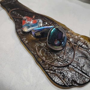 May include: A collection of dichroic glass jewelry pieces on a decorative glass tray. The jewelry includes a pendant with red and blue accents, a ring, and a necklace. The tray is brown with a leaf-like shape.