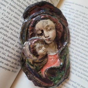 May include: A ceramic sculpture depicting a mother and child. The figures are rendered in muted tones of red, green, and brown, with delicate facial features. The artwork is set against a blurred background of a book's pages.