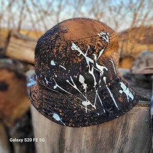 May include: A brown bucket hat with a black and white splatter paint design. The hat has a wide brim and a flat crown.