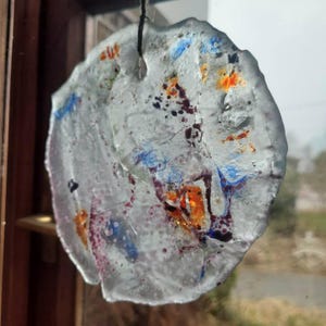 Puede incluir: Un atrapasoles de vidrio transparente de forma irregular con acentos coloridos. El atrapasoles presenta salpicaduras de tonos naranja, azul y morado. Está suspendido por un hilo negro fino, diseñado para colgar en una ventana.