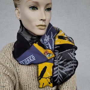 May include: A multi-colored scarf made of different fabrics with a floral pattern and the word "CLOSED" printed on it. The scarf is worn by a mannequin and has a small flower pin attached to it.