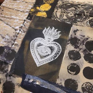 May include: A collection of fabric swatches with various patterns and textures. A black swatch features a white illustration of a heart with an eye and flames. Other swatches display script, floral designs, and circular patterns in shades of black, white, and tan.