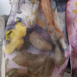 Peut inclure: Une &eacute;charpe en soie avec un motif floral dans des tons de jaune, orange, violet et marron. L'&eacute;charpe est r&eacute;alis&eacute;e avec une technique de teinture naturelle, cr&eacute;ant un design unique et artistique.