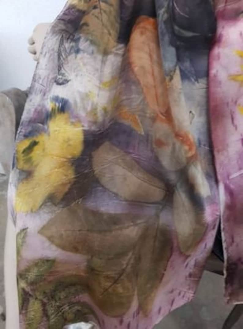 Peut inclure: Une &eacute;charpe en soie avec un motif floral dans des tons de jaune, orange, violet et marron. L'&eacute;charpe est r&eacute;alis&eacute;e avec une technique de teinture naturelle, cr&eacute;ant un design unique et artistique.