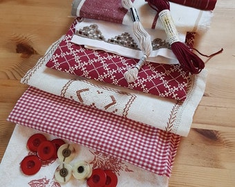 Pode incluir: Uma coleção de tecidos dobrados em tons de vermelho, branco e cinza, incluindo têxteis estampados e xadrez vichy. Dois carretéis de linha e vários botões vermelhos e brancos também são visíveis. Os itens estão dispostos em uma superfície de madeira.