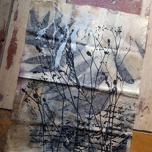 May include: A textile artwork featuring a botanical print in black and white. The design includes silhouettes of plants and leaves, with a handwritten signature at the bottom. The background is a neutral beige color, with a distressed look.