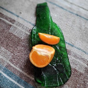 Könnte beinhalten: Ein grünes, flaschenförmiges Serviertablett aus Glas mit zwei orangefarbenen Zitrusfruchtspalten. Das Tablett ist tief smaragdgrün und hat eine strukturierte Oberfläche. Die Fruchtspalten sind leuchtend orange und weiß.