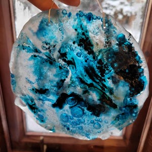 Op de afbeelding: Een ronde, doorschijnende glazen ornament met een uniek abstract ontwerp. Het ornament heeft wervelingen van blauwe en zwarte inkt, waardoor een marmer effect ontstaat. Bellen zijn verspreid over het ontwerp. Een dun touwtje is bevestigd om op te hangen.