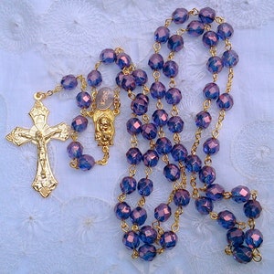 May include: A gold rosary with purple glass beads. The rosary features a gold crucifix with a figure of Jesus Christ.