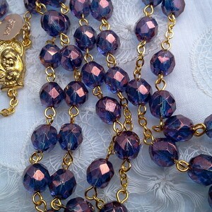 May include: A purple glass rosary with gold accents. The beads are faceted and have a metallic sheen. The rosary is strung on a gold chain.