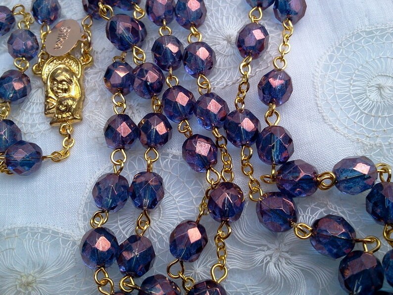 May include: A purple glass rosary with gold accents. The beads are faceted and have a metallic sheen. The rosary is strung on a gold chain.