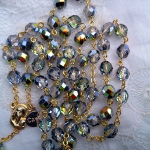 May include: A gold chain rosary with iridescent blue and green crystal beads. The centerpiece features a gold pendant with a depiction of the Virgin Mary and baby Jesus.
