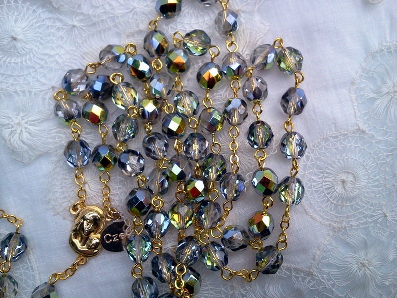 May include: A gold chain rosary with iridescent blue and green crystal beads. The centerpiece features a gold pendant with a depiction of the Virgin Mary and baby Jesus.