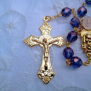 May include: A gold-toned rosary with purple glass beads and a gold crucifix featuring a figure of Jesus Christ. The rosary also includes a gold-toned pendant with a depiction of the Virgin Mary and baby Jesus.