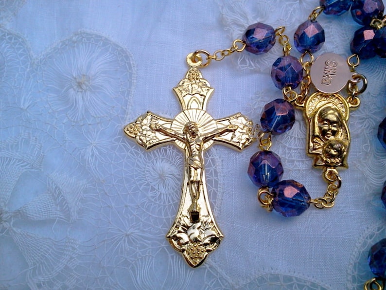 May include: A gold-toned rosary with purple glass beads and a gold crucifix featuring a figure of Jesus Christ. The rosary also includes a gold-toned pendant with a depiction of the Virgin Mary and baby Jesus.