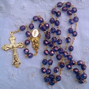 May include: A gold rosary with purple glass beads. The beads are faceted and have a metallic sheen. The rosary has a gold crucifix and a gold centerpiece with a depiction of the Virgin Mary.
