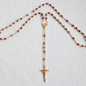 May include: A rosary featuring deep red faceted beads linked by a gold-toned metal chain. The design includes a central gold-toned metal centerpiece and a gold-toned crucifix pendant at the bottom, showcasing a detailed figure of the crucified Christ.