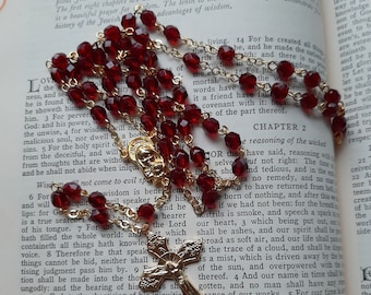 May include: A red and gold rosary with a crucifix pendant. The rosary is resting on an open book with text about wisdom and the reasoning of the wicked.