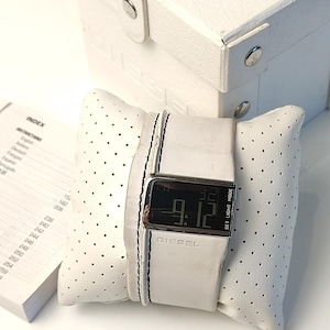 May include: White digital wristwatch with a black face and silver accents. The watch is on a white leather strap with black stitching. The watch face displays the time as 9:12. The watch is in a white box with a perforated top.