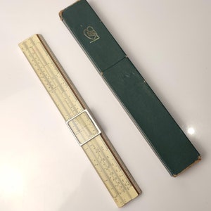 May include: A vintage slide rule with a white face and black markings in a green cardboard box. The box has the word "Faber" printed on it.